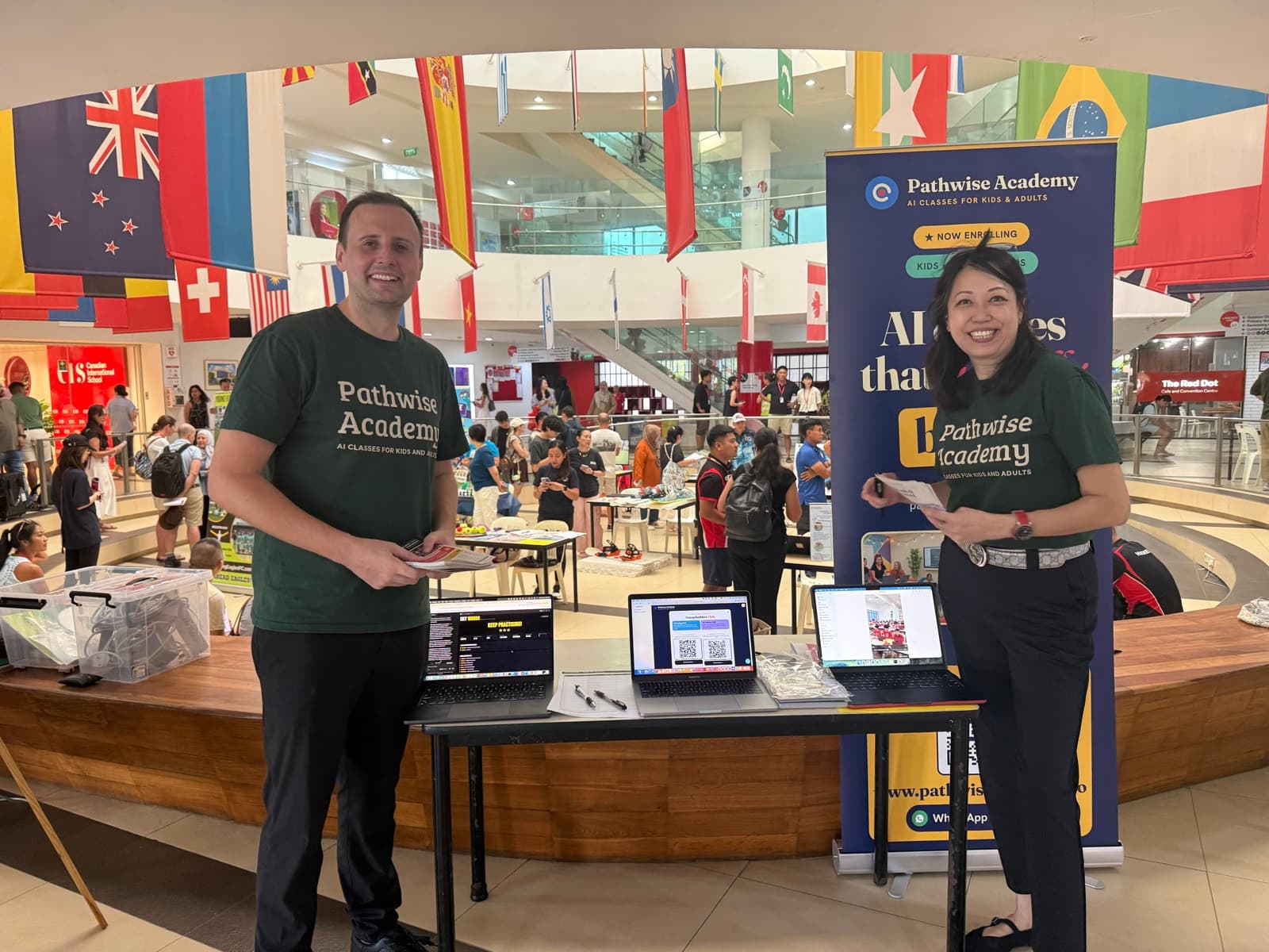 Two Pathwise Academy team members at a Pathwise stall in an international school atrium, laptops open on the table and national flags hanging overhead